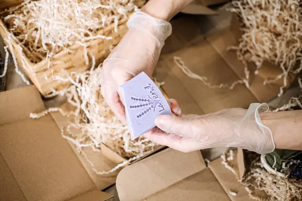 Eldivenli eller, parçalanmış kağıtlarla dolu Kraft ambalajının üzerinde lavanta sabunu tutuyor. El yapımı iş, butik markalaşma, ürün sunumu, doğal cilt bakımı. Yüksek kalite fotoğraf