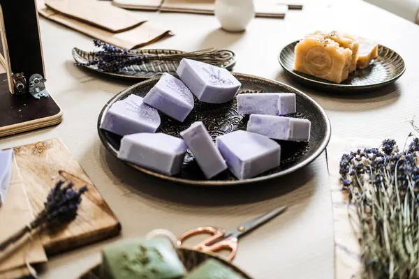 Kurutulmuş otların ve zanaatkar aletlerin yanında seramik bir tabakta sergilenen lavanta sabunları. El yapımı sabun, sanayi karşıtı güzellik, yavaş üretim, doğal cilt bakımı. Yüksek kalite fotoğraf