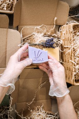 Eldivenli eller, lavantalı sabunu dilimlenmiş dolgunun ve açık kraft ambalajının üzerinde tutar. Ürün sunumu, butik markalaşma, el yapımı iş, sanatsal sabun tasarımı. Yüksek kalite fotoğraf