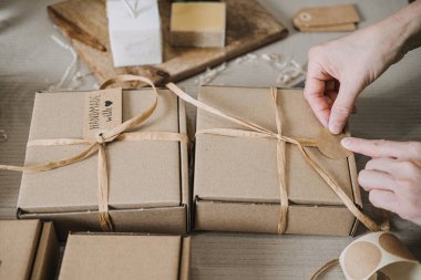 Eller Kraft hediye kutularını el yapımı sabunların ve boş etiketlerin yanında kurdele ile bağlar. Küçük seri üretim, elle ambalajlama, zanaatkar ekonomisi, yerel iş kimliği. Yüksek kalite fotoğraf