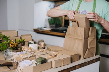 Kişi, el yapımı sabun ve çiçek dekoru dolu kraft kutularını bir kese kağıdına koyar. Eko hediyeler, plastiksiz tatiller, sanatsal hediyeler, sürdürülebilir ambalajlar. Yüksek kalite fotoğraf