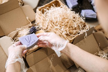 Eldivenli eller, lavantalı sabunu dilimlenmiş dolgunun ve açık kraft ambalajının üzerinde tutar. Ürün sunumu, butik markalaşma, el yapımı iş, sanatsal sabun tasarımı. Yüksek kalite fotoğraf