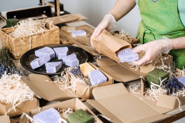 Kişi, lavanta sabununu öğütülmüş dolgu bezi ve el yapımı çubuklarla çevrili bir kese kağıdına koyar. El yapımı ürün nakliyesi, bağımsız güzellik sevkiyatı, el işi markası lojistiği, eko-paketleme iş akışı. 