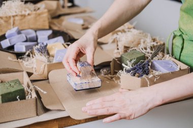 Kişi, el yapımı sabunlar, kurutulmuş lavanta ve çevre dostu dolgu malzemelerinin yanına kraft ambalajı yapıştırıyor. Üreticiler için görsel kimlik, yavaş marka değerleri, minimalist ambalajlama, sanatsal tasarım. Yüksek kalite fotoğraf