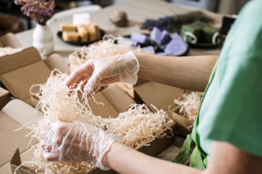 Eldivenli eller, kraft ambalajında kurumuş çiçeklerle yeşil ve lavanta sabunları düzenler. El yapımı iş, çevre dostu ambalaj, doğal cilt bakımı, sanatsal sunum. Yüksek kalite fotoğraf