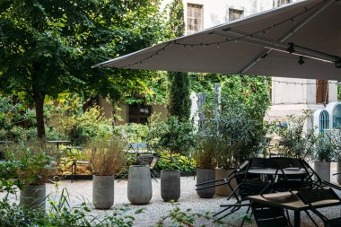 Açık hava bahçe kafesi, büyük gölge şemsiyesi, saksı bitkileri, ve yemyeşil bir avluda siyah sandalyeler. Sürdürülebilirlik, şehir yeşilliği, sosyal alanlar, rahatlama. Yüksek kalite fotoğraf