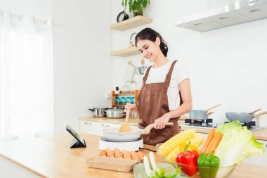 Gülümseyen genç ve güzel bir kadın mutfakta vejetaryen yemekleri pişiriyor genç bir bayan yemek için yemek pişiriyor ve mutfakta dijital tabletten yemek tarifleri alıyor. Kendi başına yemek yapmayı öğreniyor.