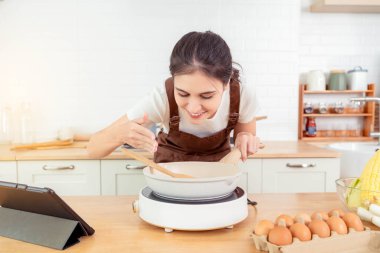 Mutfakta elektrikli ocağın yanında duran kadın, anne yemek pişiriyor ve tavadaki lezzetli yemeğin güzel kokularını kokluyor. Güzel kokular, diyet öğle yemeği kıyafetlerini içeride, modern mutfak önlüğüyle pişirmeye devam ediyor.