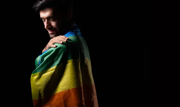 LGBTQ Handsome man looking at rainbow flag or gay pride flag He is Gay Pride and Proud himself Gay hugging himself and touching rainbow flag softly with black background LGBTQIAN+ Pride Month concept