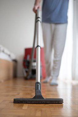 Unrecognizable blurry out of focus male, using a vacuum cleaner brush and extension rod on laminated wooden tile surface inside a generic apartment. Close, upwards looking shot, shallow depth of field.