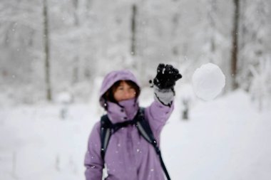 Tanımlanamayan genç, beyaz bir kadın, kartopu atıyor. Kar yağan orman sahnesi, seçici odak, başlık kafaya.