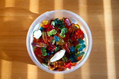 Plastik yuvarlak kabın içinde çeşitli renkli jelibon şekerler. Tahta bir masaya kurulmuş, güneş ışığıyla aydınlatılmış, yakın çekim, insan yok..