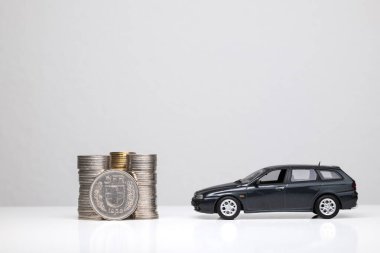 Small metal toy model car next to a stack of coins and a 5 Swiss Francs coin isolated on white. Close up shot, side profile, dark gray station wagon, no people clipart