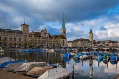 24-12-2024 Zürih, İsviçre. Limmat nehri manzaralı Fraumunster kilisesi ve köprüsü. Kısmen güneşli bir kış günü, su yansımaları, köprüden geçen turist..