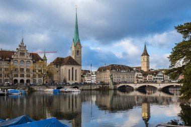 24-12-2024 Zürih, İsviçre. Limmat nehri manzaralı Fraumunster kilisesi ve köprüsü. Kısmen güneşli bir kış günü, su yansımaları, köprüden geçen turist..