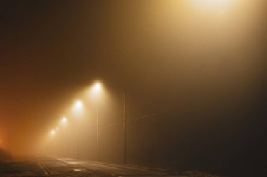 Kasvetli turuncu sokak ışıkları sisli bir kış gecesinde boş şehir yolunu aydınlatıyor. Araba yok, insan yok.