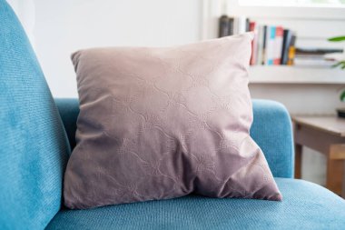 Açık mavi kumaş kanepenin köşesinde şişirilmiş yastık. Geniş açı açısını kapat, yüzeysel alan derinliği, doğal pencere ışığı, arka planda bulanık kitap rafı, insan yok.