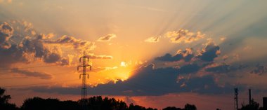 Gün batımında bir tarlada yüksek voltajlı elektrik direği. Düşük, geniş açı, dramatik günbatımı bulutları, insan yok.