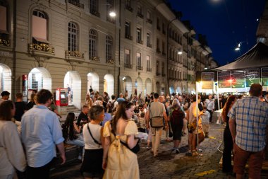 07-08-2025 Bern, İsviçre. Kalabalık sokak yemeklerinin ve Buskers sokak müzik festivalinin tadını çıkarıyor. Mavi saat ışığı, uzun pozlama, bulanık insanlar, geniş açı.