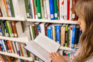 Kafkas, uzun, sarışın kadın, kütüphanenin içinde kitap raflarının yanında kitap okuyor..