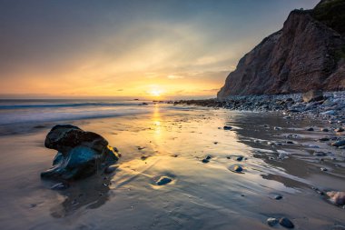 Altın güneş ışığı, Dana Point 'in kayalıklarının yakınındaki dalgalar yavaşça kıyıya vururken ıslak kumu yansıtıyor ve sakin bir sahil atmosferi yaratıyor..