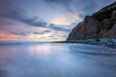 Güneş Dana Cove 'un uçurumlarının ardında batarken dalgalar yavaşça kıyıya vurur ve gökyüzünü yumuşak pastellerle boyarlar..