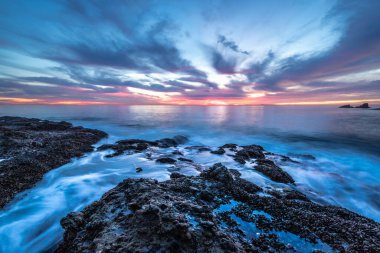 Güneş sakin suların ve Crescent Bay Beach, Güney Kaliforniya 'nın kayalık kıyı şeridinin üzerinde batarken gökyüzünü canlı renkler kaplıyor..