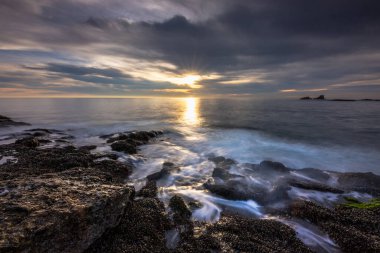 Soft waves lap against rocky shores as a vibrant sunset casts a warm glow over the tranquil waters of Crescent Bay in Laguna Beach, California. clipart