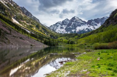 Görkemli Maroon Çanları, yemyeşil yemyeşil ve engebeli dağlarla çevrili Maroon Gölü 'nü Aspen, Colorado' da kasvetli bir gökyüzü altında yansıtıyor..