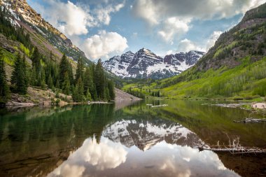 Göz kamaştırıcı Maroon Bells dağları berrak bir göl üzerinde yükselir, yemyeşil ve parlak mavi bir gökyüzü altında karla kaplı zirveler.