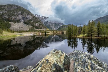 Clear waters of Officers Gulch Pond mirror the stunning Rocky Mountains under a cloudy sky, showcasing serene summer nature in Frisco. clipart