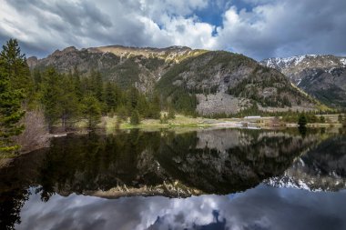 Clear waters of Officers Gulch Pond mirror the stunning Rocky Mountains under a cloudy sky, showcasing serene summer nature in Frisco. clipart