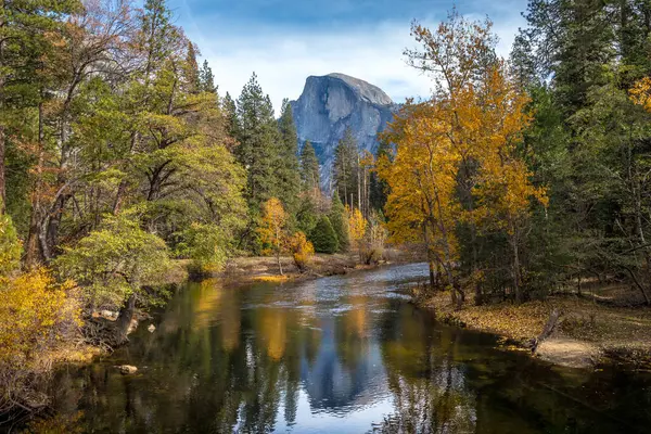 Bu pitoresk manzara, Yosemite 'deki canlı sonbahar yaprakları ve dingin dağların çerçevelediği sakin bir nehre karşı Half Dome' u sergiliyor..