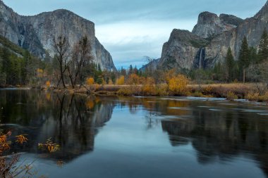 Sonbahar yaprakları Merced Nehri 'nin sakin sularını yansıtıyor. Gün batımında Yosemite Vadisi üzerinde yükselen dağlar, dingin bir atmosfer yaratıyor..