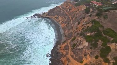 Bluff Cove, Palos Verdes Gayrimenkul 'ün sinematik hava gösterimi, bulutlu bir günde dalgalar engebeli kıyı şeridine çarpıyor.