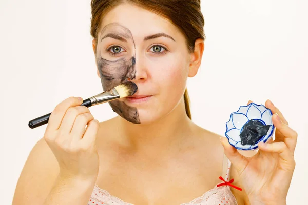 女人在她的脸上涂上黏土碳酸脱毒面罩 雌性用黑色化妆品和刷子托着碗 美容美发吓吓 — 图库照片