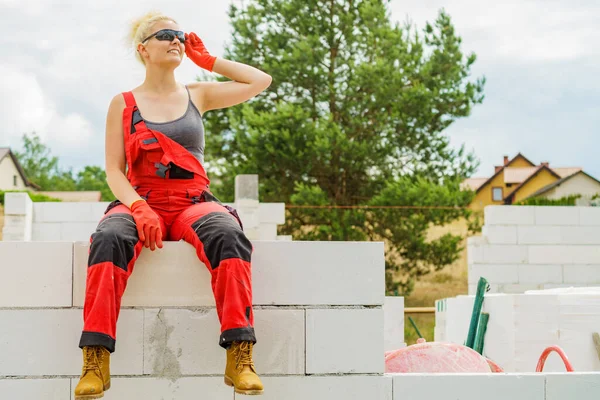 建設現場で作業着を身に着けている女性 女性は仕事を休み 壁に座っている 部分的に新しい家の初期の段階を構築しました — ストック写真