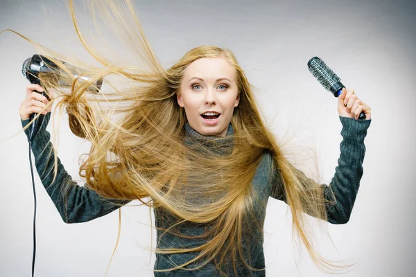 Female with blowing messy hair holding accessories hairdryer and brush. Blonde woman styling her very long disobedient hair.