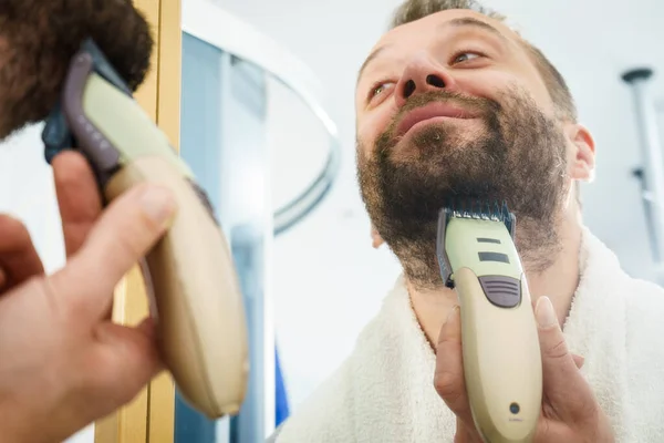 Bearded man looking at himself in mirror trimmng, shaving his beard using electric timmer razor.