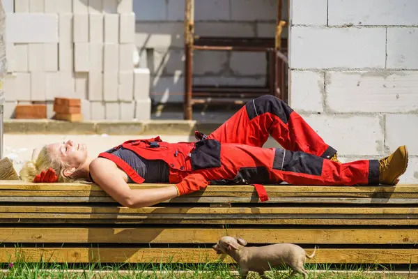 Woman in dungarees relaxing after hard work on construction site. Young female lying outdoor taking a break