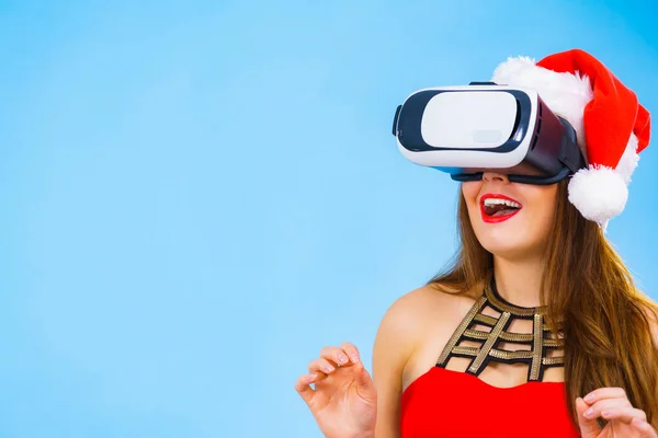 Woman wearing santa claus hat exploring space with virtual reality goggles headset. Amazed girl watching 3d film tour in vr glasses box. New generation cyber christmas concept, on blue.