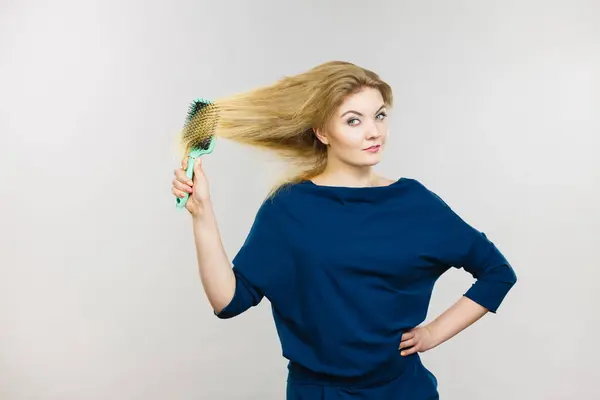 Woman combing her hair with brush. Young female with beautiful natural blond straight long hairs, studio shot on grey