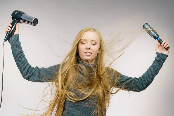 Female with blowing messy hair holding accessories hairdryer and brush. Blonde woman styling her very long disobedient hair.