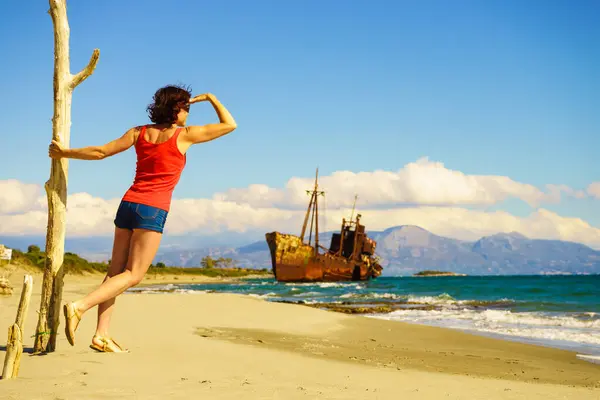 旅行自由 海滩上成熟的女游客享受暑假 一艘废弃的旧船在后面失事的船 图库照片
