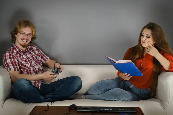 坐在沙发上消磨时间的情侣 玩电子游戏的人和女性阅读书 免版税图库图片