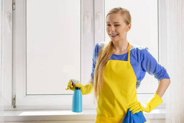 穿着黄色手套的年轻女子在家里用抹布和喷雾清洁剂擦拭窗玻璃 清洗概念 图库图片