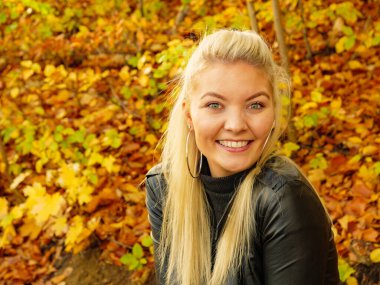 Blonde woman wearing fashionable outfit. Happy female in black leather ramones jacket walking in autumnal park, enjoying sunny day clipart