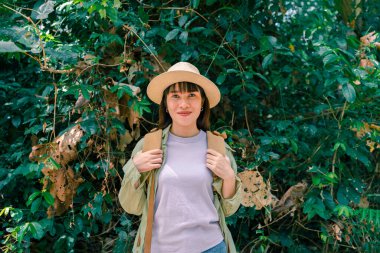 Ormanda sırt çantası ve hasır şapkalı bir kadının portresi, yürüyüş yapan gülümseyen bir kız, yaz yürüyüşü yapan, dağ yolunda yürümekten zevk alan mutlu bir kadın.