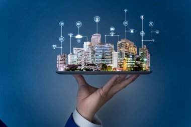 Businessman hand holding tablet then land, trees and tall buildings on it, selling real estate online. Devices connected to digital storage in data center via internet. IOT. Smart home.