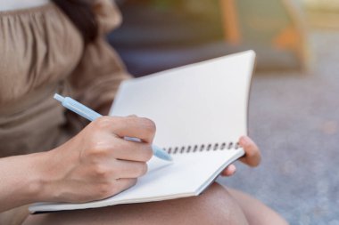 Note Taking Concept. Side view close up portrait of attractive asian woman writing in her diary, copy space. In the morning a women sitting and writing on note book in public park.